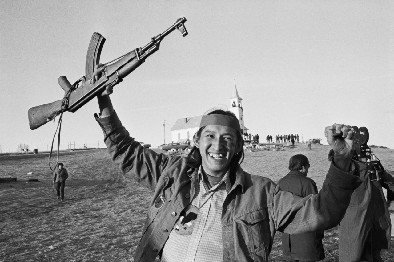 Black and white photograph of Robert Onco holding a rifle above his head at the Wounded Knee occupation. The church is in the background with a group of AIM supporters standing outside.