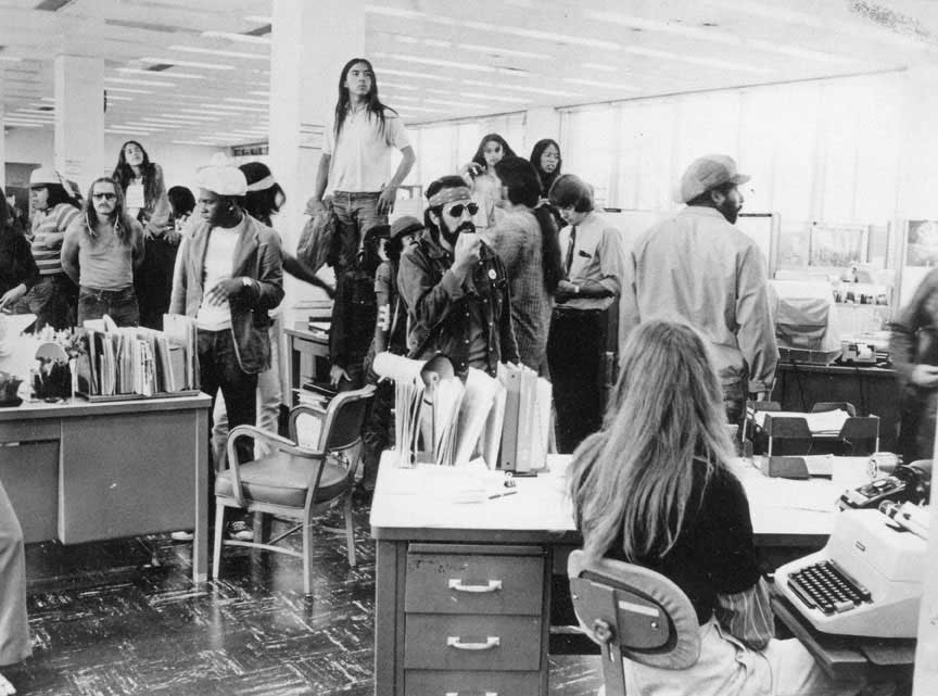 Black and white photograph of sixteen Black, Native, and White men and women protestors inside an office in the Bureau of Indian Affairs. Some of the BIA employees are also in the room.