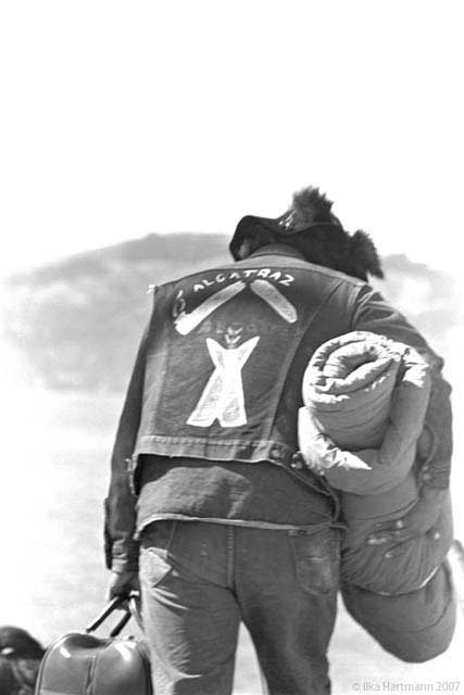 Black and white photograph of AIM member/supporter holding a suitcase and sleeping bag. He is photographed from the rear, walking away from the viewer. On the back of his jacket the painted words ”Alcatraz”, a smoking pipe split in two pieces, and a figure holding her arms up, are displayed.