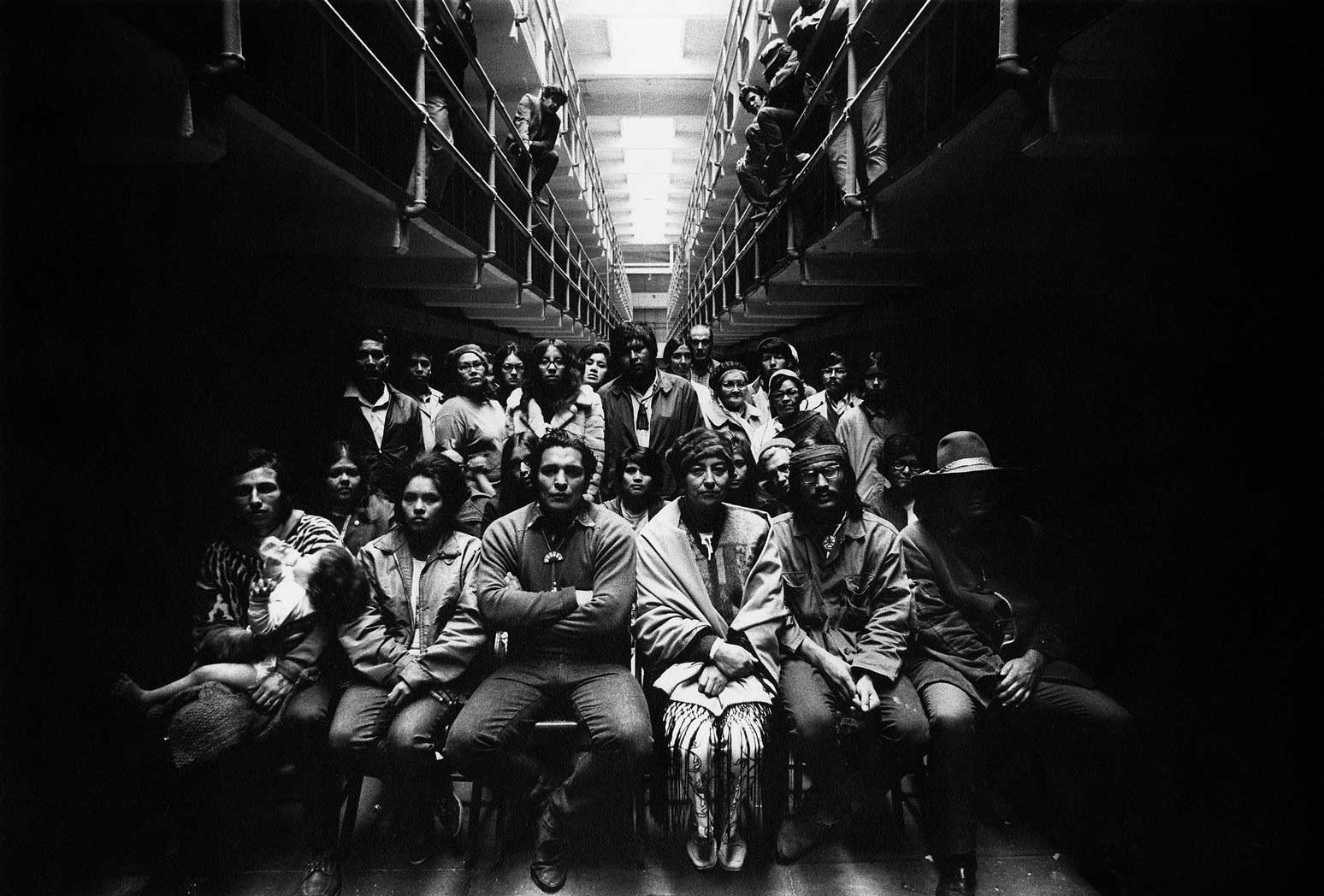 Black and white photograph of a large group of AIM members, men and women, are directly facing the camera. They are seated in a cell block indoors, at Alcatraz Prison with Richard Oakes in the middle of the front row. The group are dramatically lit from above (ambient/natural light). Some members are standing along the second row of cell blocks looking down from in the balcony above.