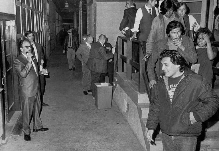 Black and white photograph of group of AIM members being escorted out of a building.  They descend a staircase at night. White men in suits and a policeman stand watching, holding flashlights as AIM members leave the building.