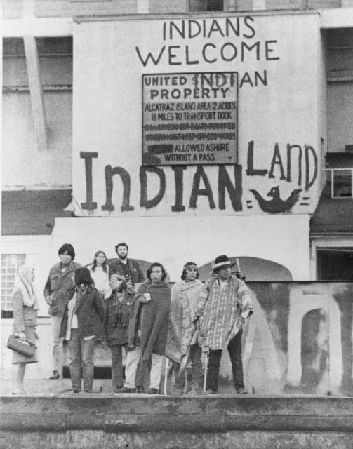 Black and white photograph of AIM members/supporters posing in front of a building with ”Indians Welcome Indian Land”.