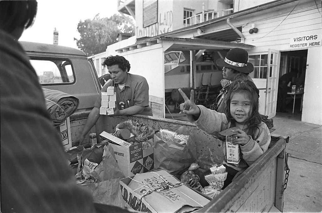 Black and white photograph of a young Yvonne Oaks making the peace sign and smiling at the camera, sitting in the back of a truck filled with supplies. Richard Oakes unloads supplies from the truck as two AIM members/supporters appear to be speaking with him.