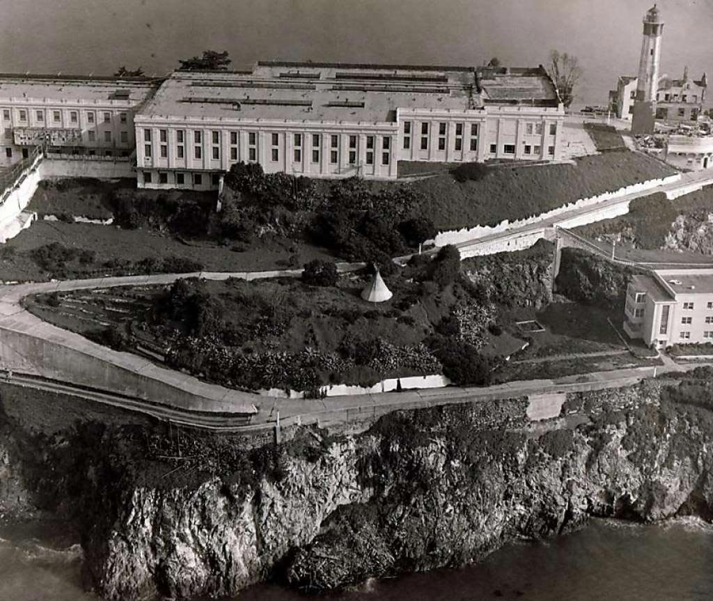 Black and white aerial photograph of tipi set up on the  grass on Alcatraz. The tipi is centered in the image and institutional buildings, the edge of the island, and water below are visible in the photograph.
