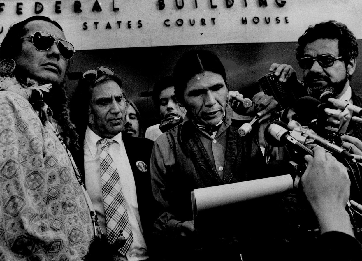 Black and white photograph showing a large group at a press conference. There are four men in the foreground. Dennis Banks appears at the center of the image reading a statement to reporters after all charges against him and Russell Means, who is the furthest on the left, were dismissed by U.S. District Judge Fred Nichol on September 17. 1974. Their attorneys, William Kunstler (second from left) and Mark Lane (right) accompany them. Photo by Charles Bjorgen/Minneapolis Star Tribune/TNS.