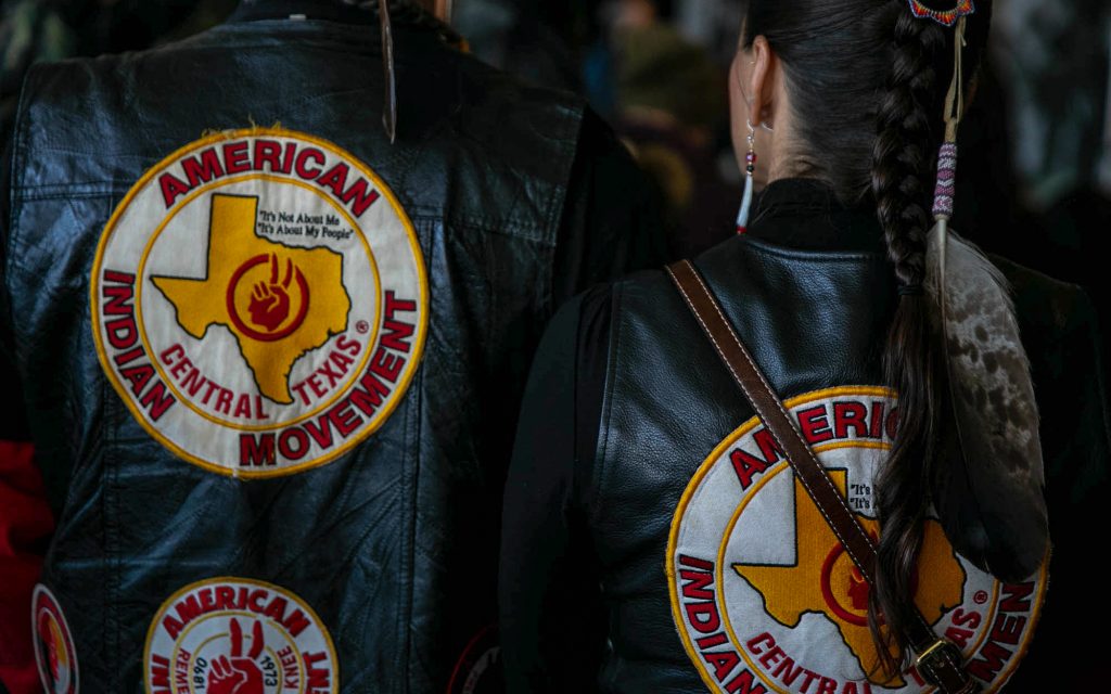 Color photograph showing the backs of two Native Americans wearing black leather jackets with a circular logo for the AIM Central Texas chapter. Logo consists of one white circle enclosing another circle with a yellow border around each circle. 'AMERICAN INDIAN MOVEMENT' stitched in red along the perimeter of the smaller circle. The center circle has the state of Texas in yellow with the red AIM logo at the center. The motto appears in black to the right above Texas and reads 'It’s Not About Me / It’s About My People'. 'Central Texas' is stitched in red beneath Texas.