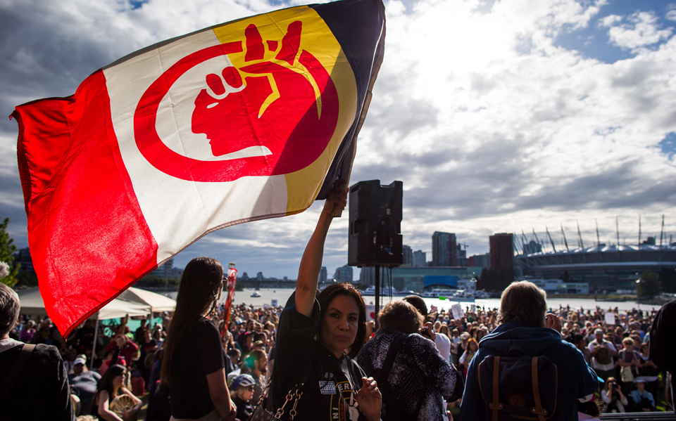 Color photograph of a large crowd at a protest against the Kinder Morgan Trans-Mountain pipeline expansion in Vancouver, British Columbia during 2018. A Native American woman appears in the center foreground of the image raising the AIM flag in her proper right hand. Photo by Darryl Dyck / The Canadian Press via AP.