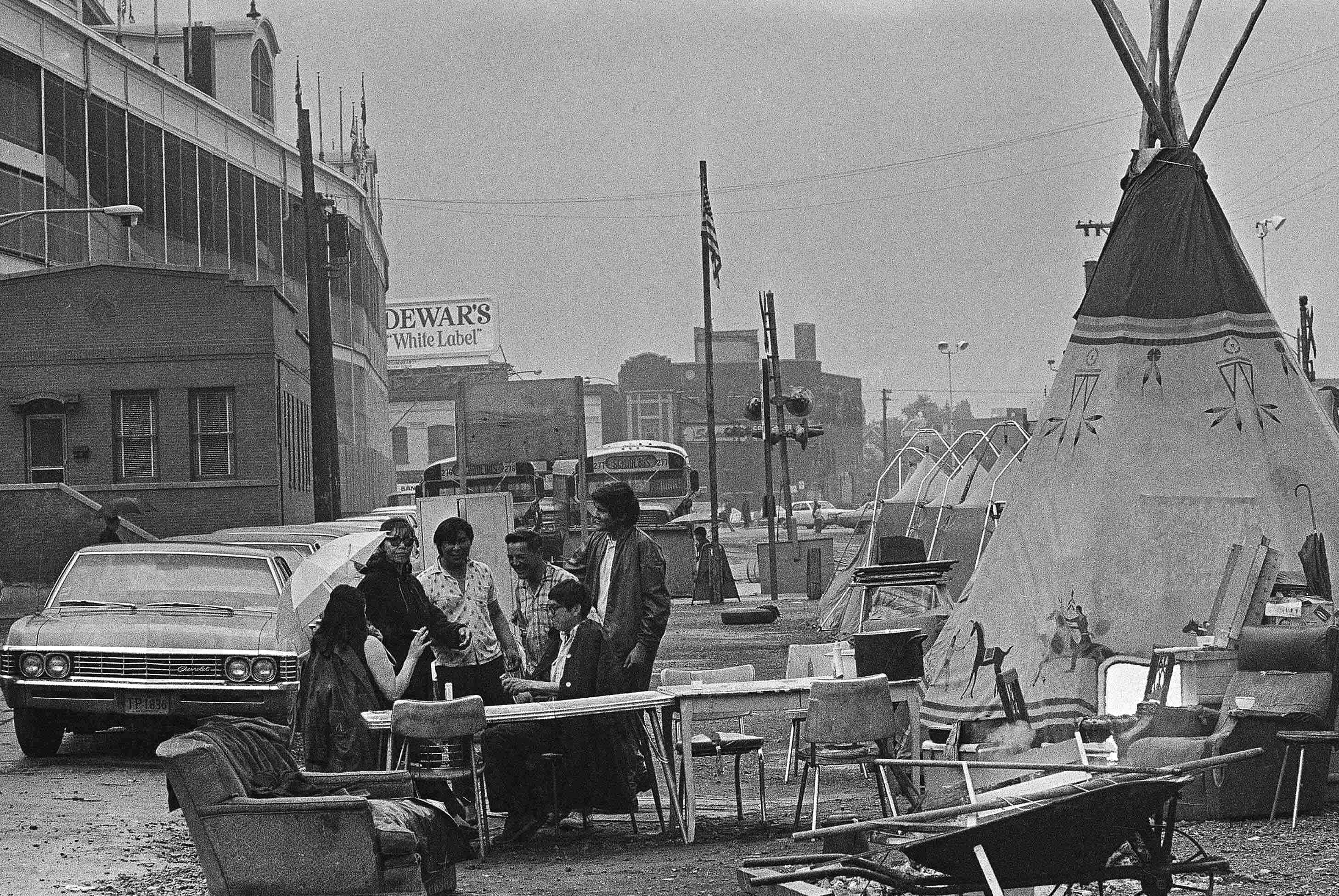 Black and white photograph of a 1970s era Chicago street scene in which two women and four men are gathered around tables and chairs; a large tipi set up near them. Cars and buses are parked nearby. An advertisement from 'Dewar’s White label' is visible on the top of a building in the distance.