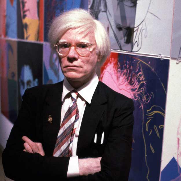 Color photograph half-length portrait of artist Andy Warhol with his arms folded in front of his work.