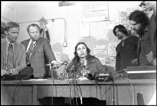 Black and white photograph of John Trudell sitting at a desk with multiple microphones for a press conference; also present are three supporters of the occupation. Two White men, dressed in suits, possible news reporters are also present.