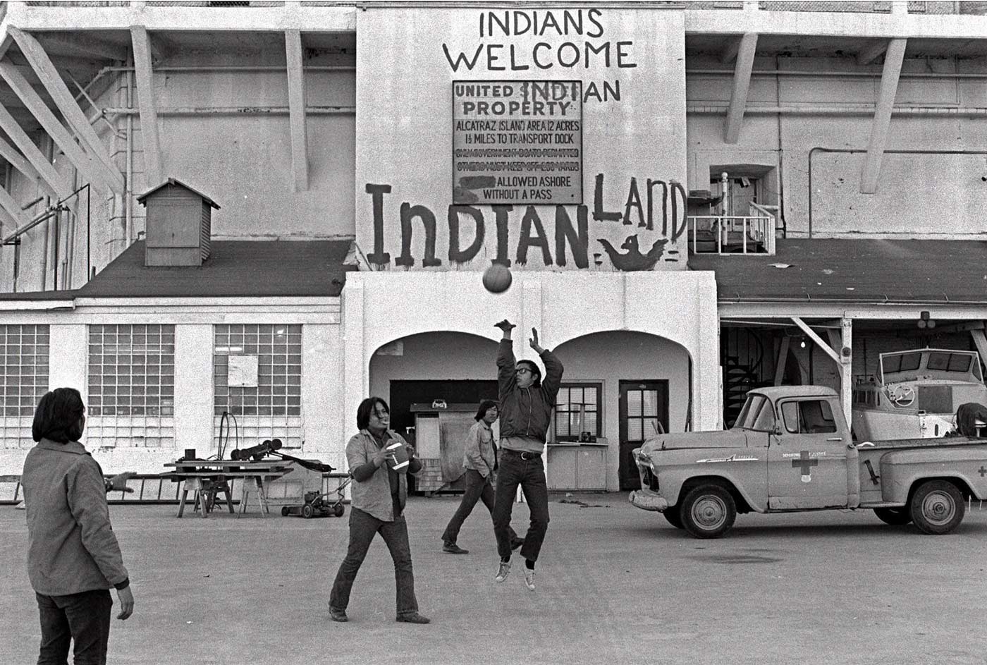 Black and white photograph of AIM members/supporters playing basketball in front of a building with ”Indians Welcome Indian Land” painted above the door/original sign.