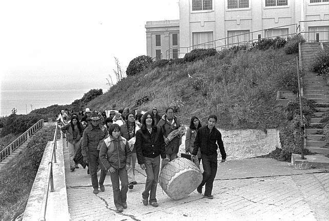 Black and white photograph of AIM members/supporters walking on Alcatraz, carrying a large drum.