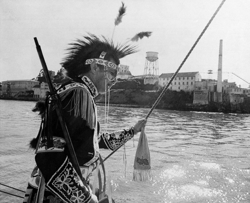 Black and white photograph of Native American man wearing traditional ceremonial clothing, Alcatraz in background.