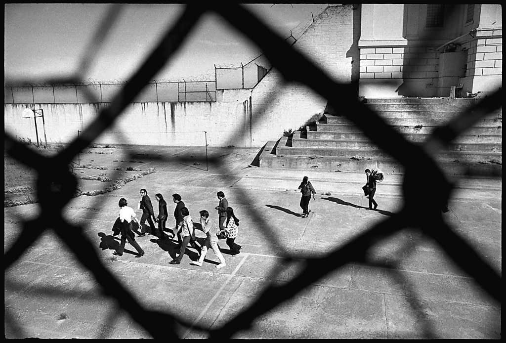Black and white photograph of AIM members/supporters walking on Alcatraz, viewed through a chain link fence.