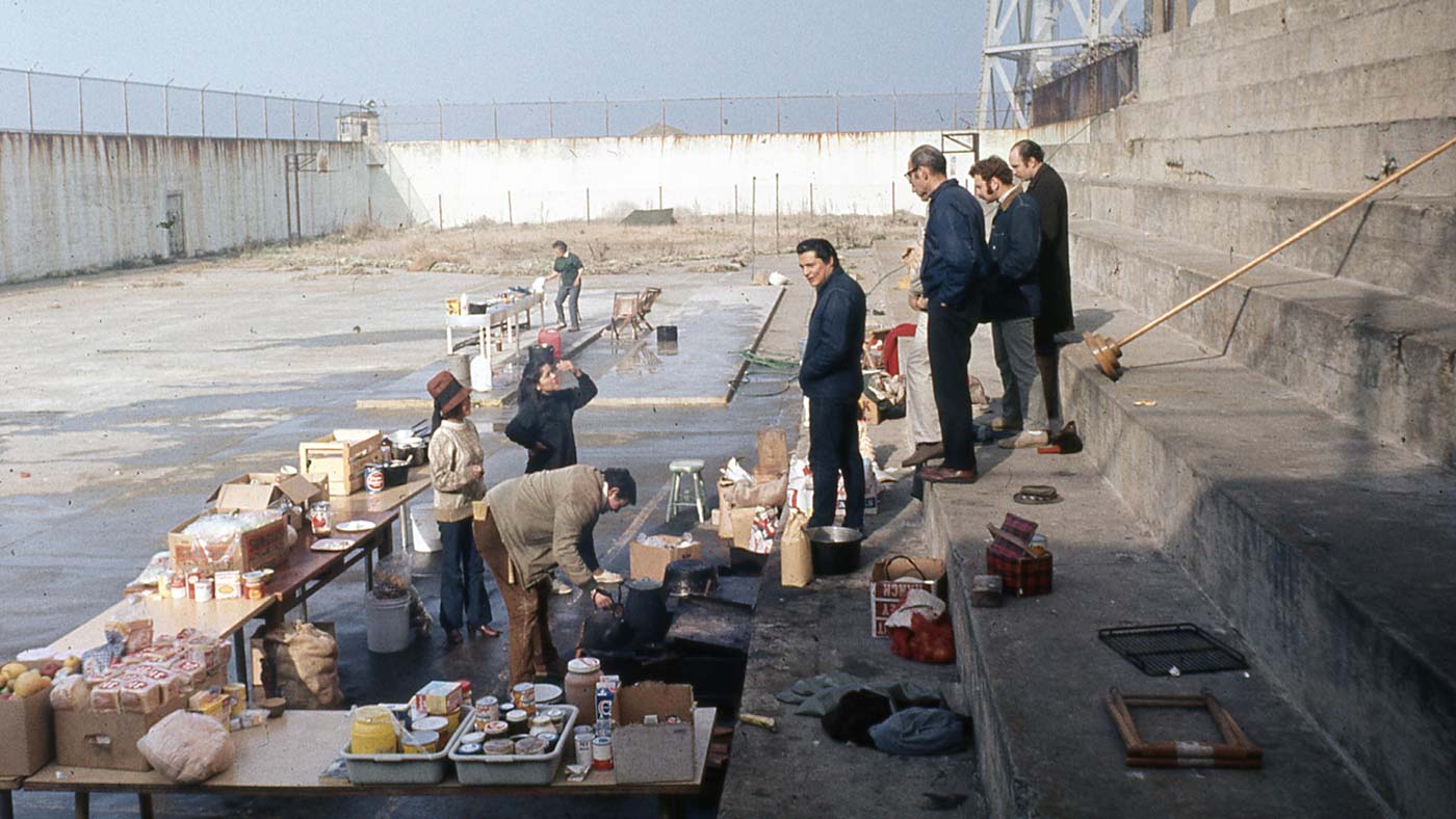 Color photograph of Richard Oakes and other AIM members/supporters surveying food and supplies laid out on tables, outdoors, on Alcatraz.