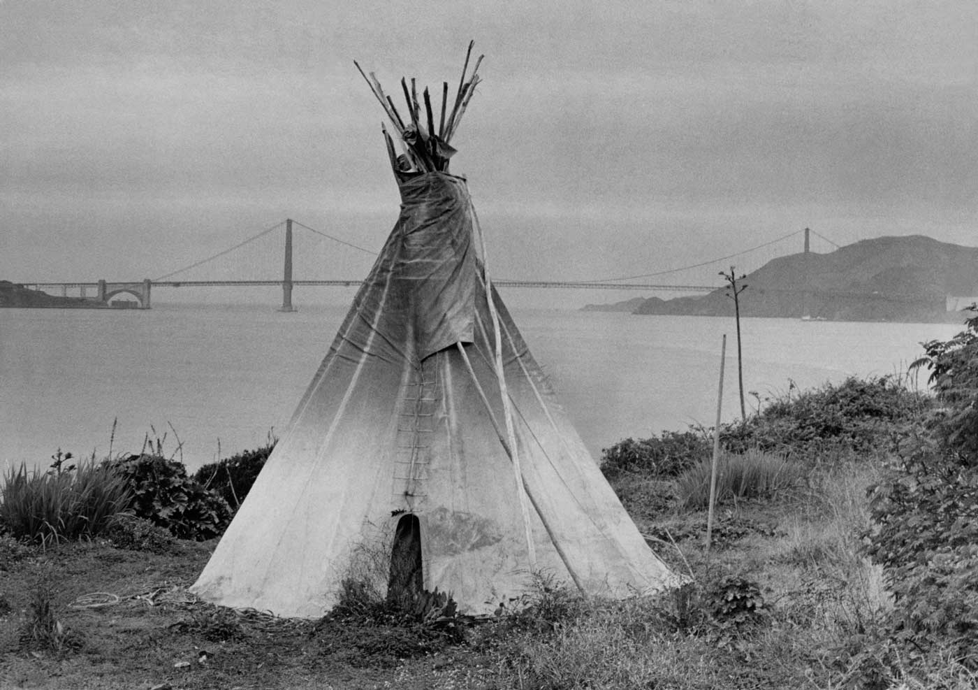 Black and white photograph of a tipi set up in the grass on Alcatraz, Golden Gate bridge and San Francisco Bay are visible in the background.