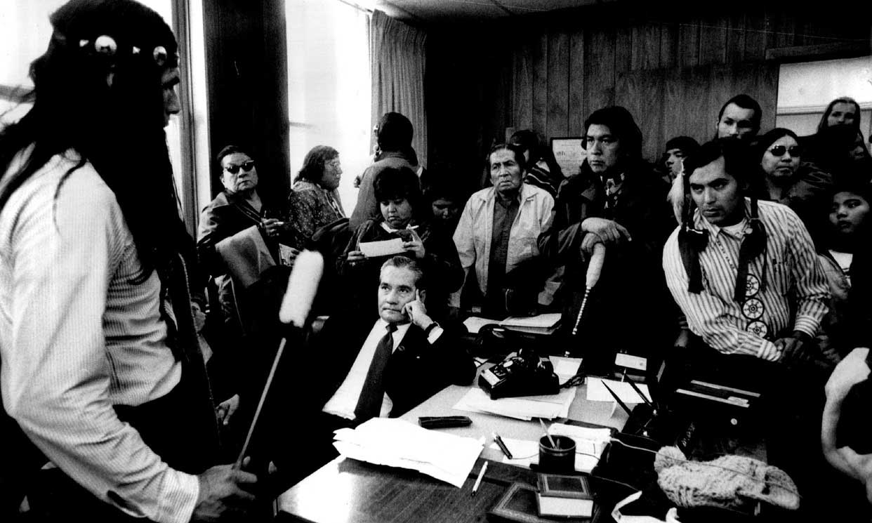 Black and white photograph of Russell Means and AMI supporters in the office of a government official. Official is seated at his desk.