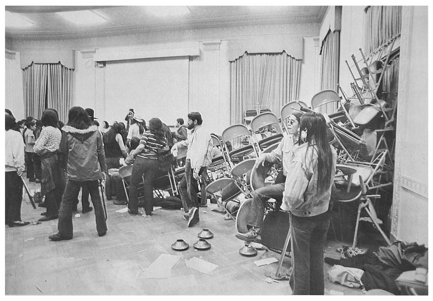 Black and white photograph of protestors inside the Bureau of Indian Affairs. Most of the protestors in the photograph are gathered on the left side of the photograph. On the right side of the photograph the chairs and other furnishings have been thrown into a large pile.