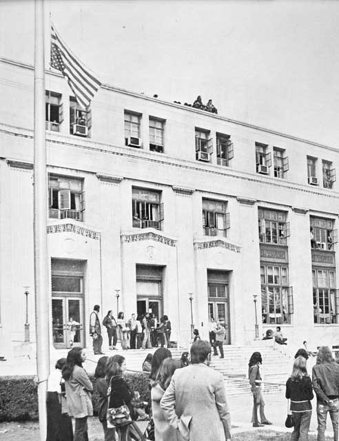 Black and white photograph showing a large crowd of men and women gathered in front of the Bureau of Indian Affairs in Washington, D.C., including on the roof of the building. The American flag is flying upside-down on the flagpole.