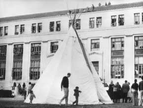 Black and white photograph of a man and two small children walking around a tipi in front of the Bureau of Indian Affairs in Washington, D.C. during the AIM sit-in at the government office. A crowd of people is standing between the tipi and the building, and eight people are sitting on the roof.