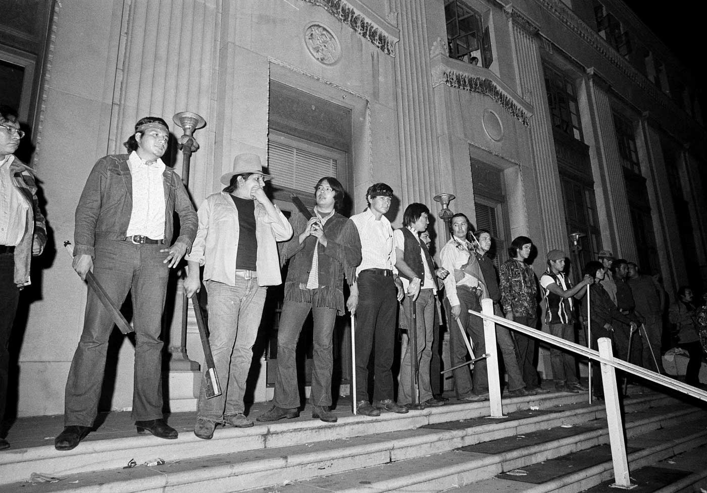 Black and white photograph of seventeen AIM men standing in front of the Bureau of Indian Affairs in Washington, D.C. Photo was taken at night and several of the men are holding clubs and sticks.