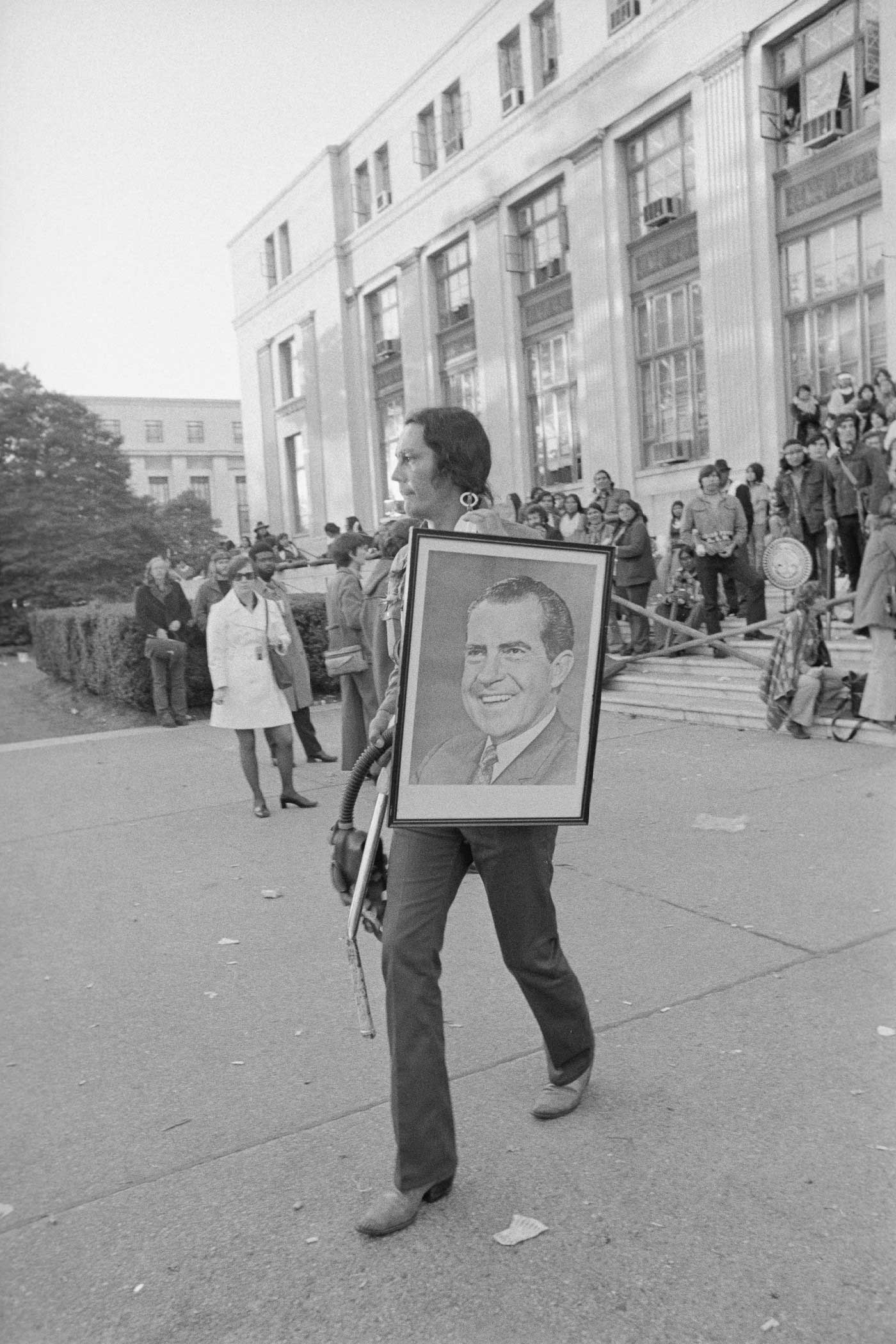 Black and white photograph of Russell Means walking out of the Bureau of Indian Affairs carrying a framed photograph of President Richard Nixon.