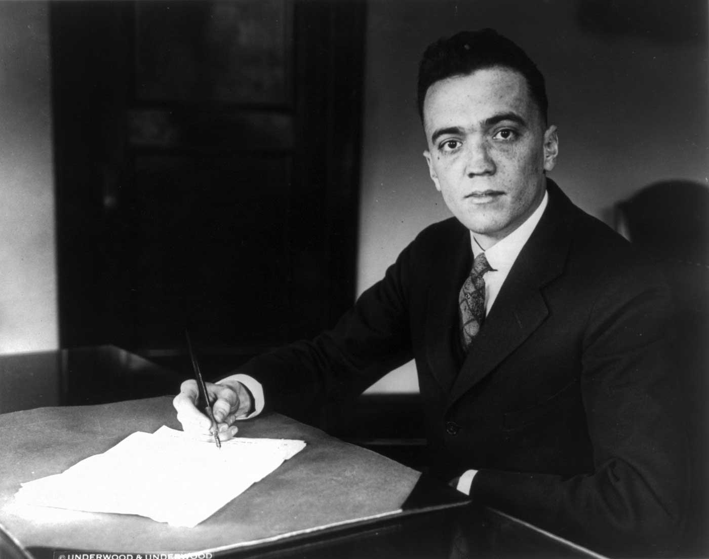Black and white photograph of a young J. Edgar Hoover, Director of the FBI from 1924-1972, sitting at a desk holding a pen above a sheet of paper. The photograph was taken on May 16, 1932.