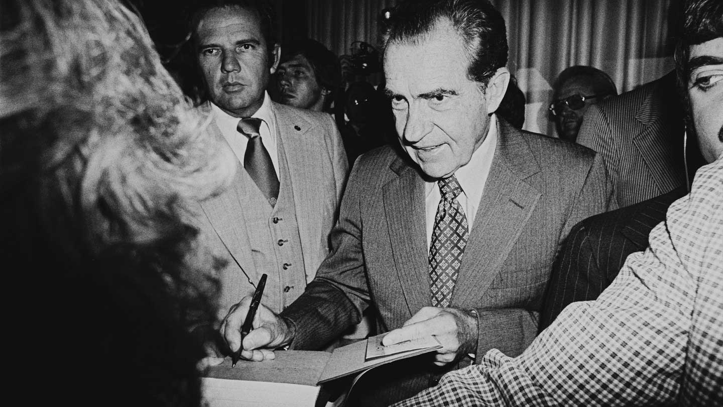 Black and white photograph of President Richard Nixon autographing a book. There is a crowd of men and women close to the President.