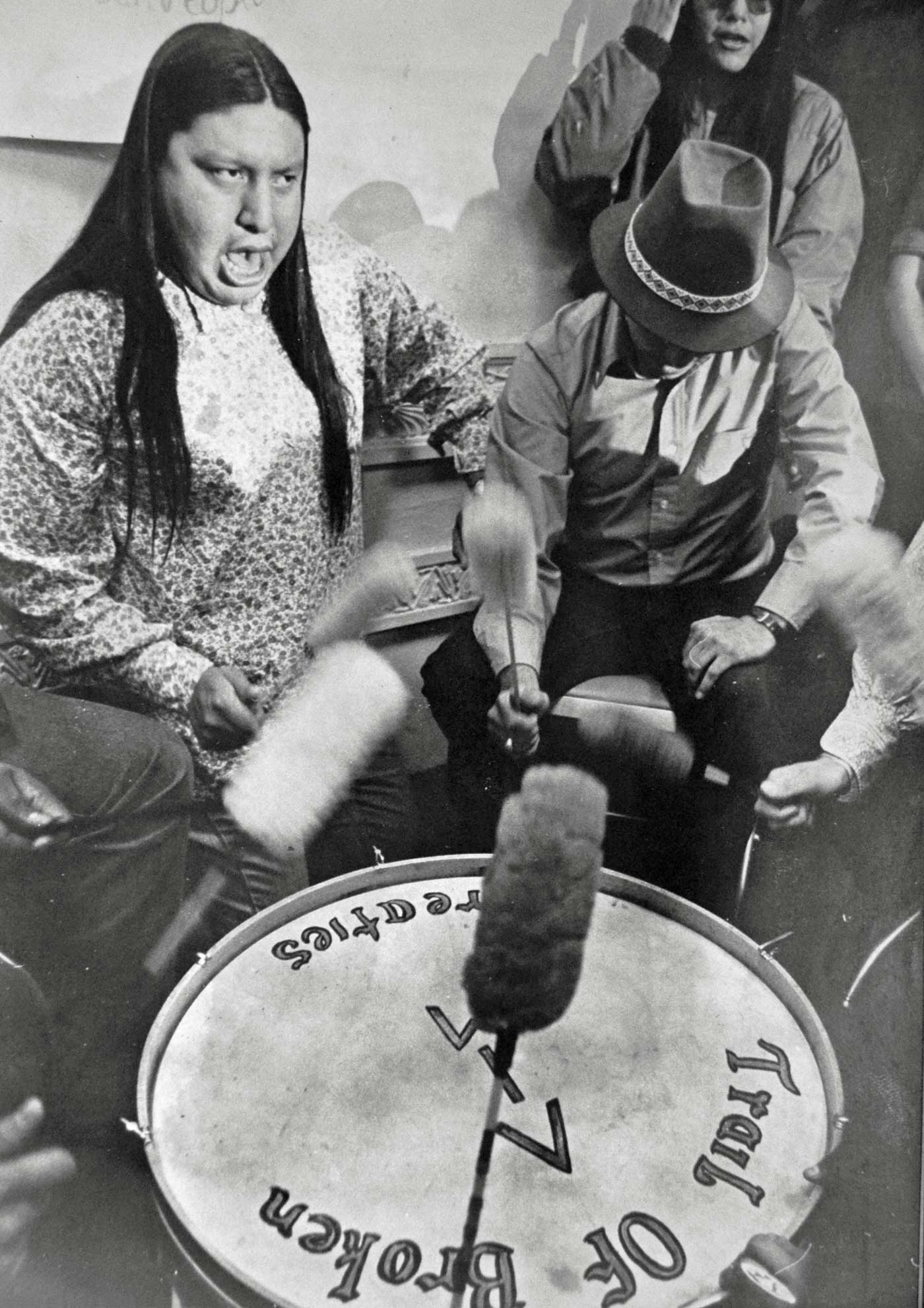 Black and white photograph of a drumming session in the Bureau of Indian Affairs in Washington, D.C. on the first day of demonstrations. ”Trail of Broken Treaties” and ”AIM” are painted on the drum.
