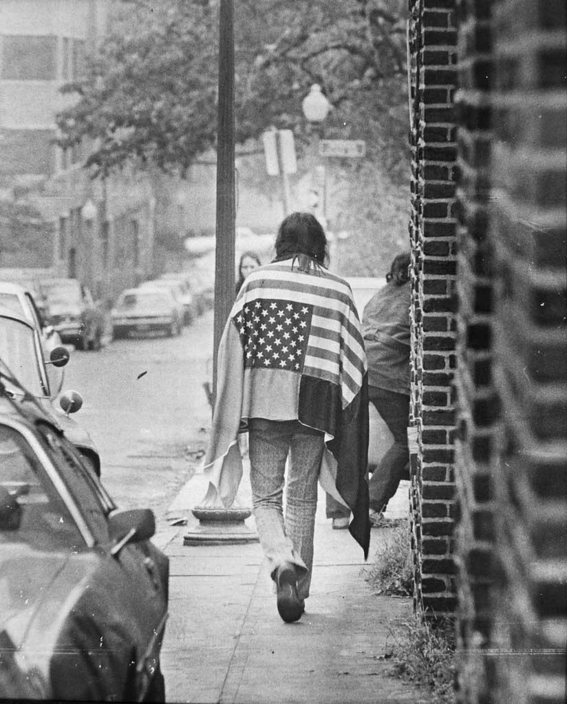 Black and white photograph of a man walking down an urban sidewalk away from the camera wearing a U.S. flag sewn onto a larger cloth, draped across his shoulders.