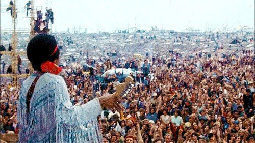 Color photograph of Jimi Hendrix performing while wearing a headband and jacket with long fringe. Hendrix is photographed from behind and a large crowd fills the background of the image.