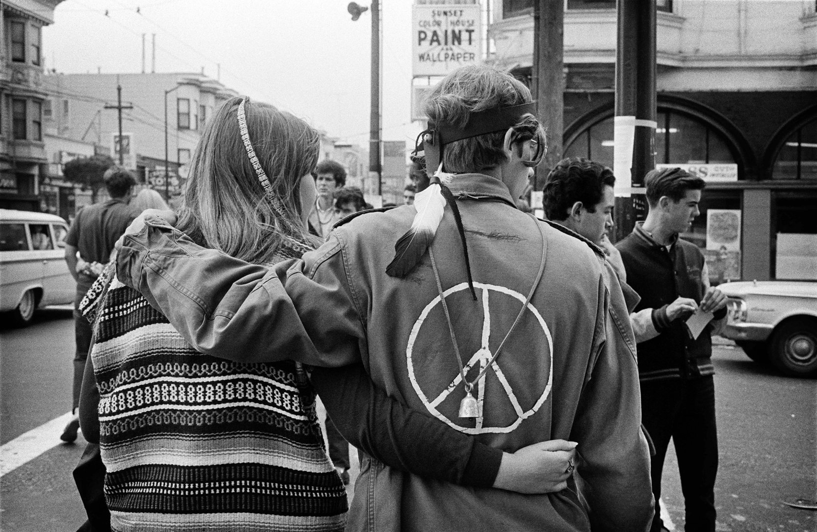 Black and white photograph showing a young White woman and man from behind. The couple has their, arms around one another and they are walking in the street. The woman wears an intricately patterned sweater and woven hair accessory. The man wears clothing that is of Native American design along with military jacket with a peace symbol drawn on the back. A group of eight young men and a woman are visible in the background. The text 'SUNSET COLOR HOUSE PAINT AND WALLPAPER' is visible on a sign above the street on a corner building.
