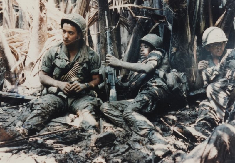 Color photograph of three Native American U.S. Army soldiers reclining on the ground surrounded by vegetation in SE Asia. They wear green uniforms, helmets, bullet straps hang around their necks. One soldier holds a rifle.