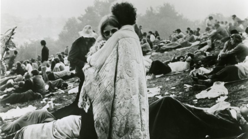 Black and white photograph taken at the Woodstock music festival in 1969. A White man and woman embrace standing, surrounded by a blanket that the man is wearing. In the background: many others sit and lay in the field, wrapped in blankets, facing toward the left of the photo.
