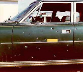 Color photograph of the driver’s side door of a green car riddled with bullet holes.