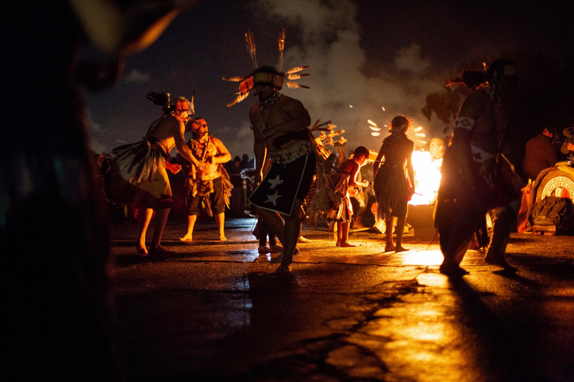 Color photograph of a group of dancers in Native American ceremonial apparel dancing by the light of a fire at dawn on Alcatraz Island. Photo by Talia Herman/The Guardian.