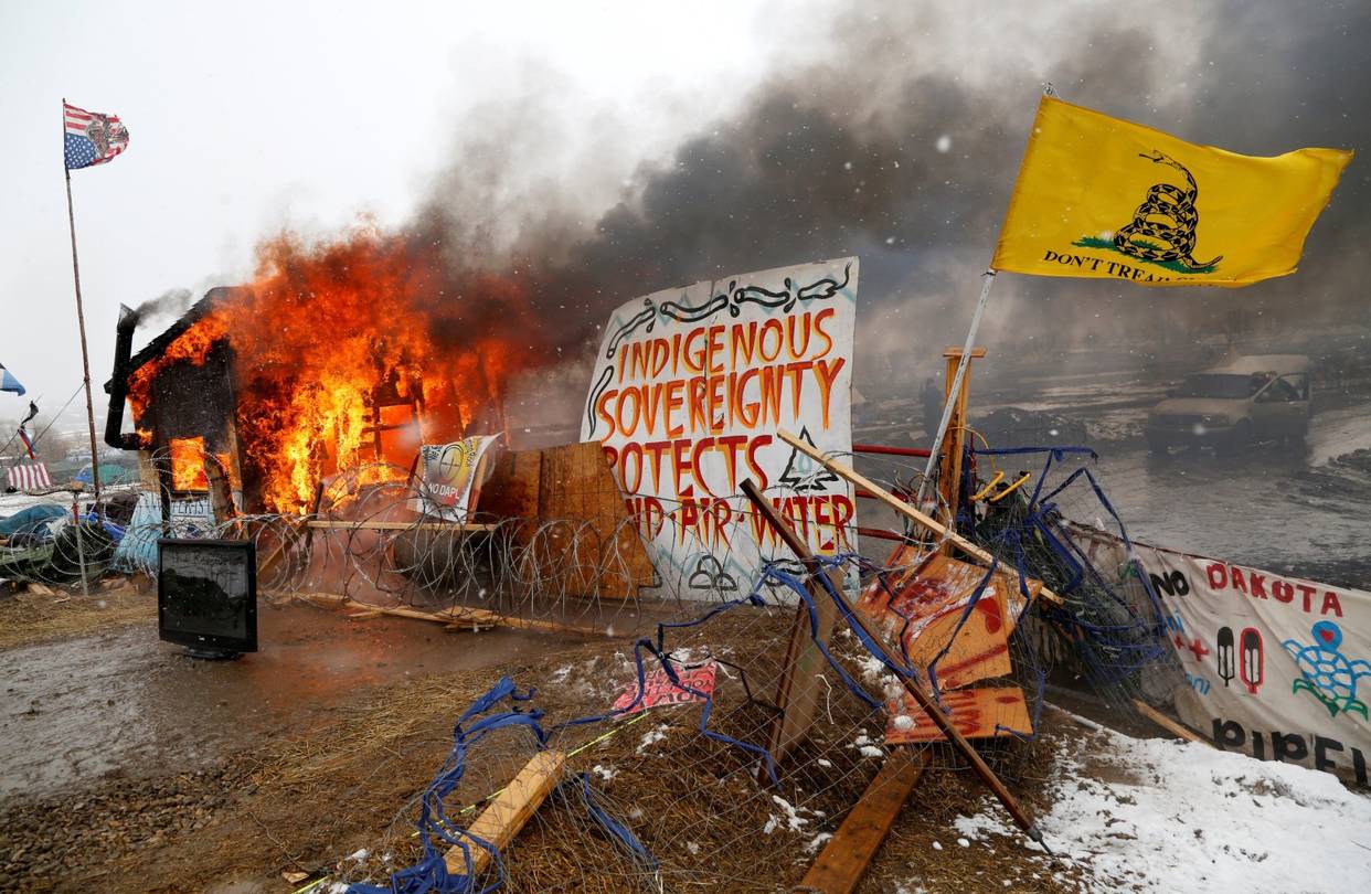 Color photograph showing the cleared protest site for activists against the Dakota Access Pipeline after police pressed them to leave. As protestors left the site, some set fire to camp structures as part of a leaving ceremony. Barbed wire and fencing adorned in protest signs and supporting an upside-down American flag and the yellow 'Don’t Tread on Me' flag is collapsing in the foreground. One sign at the center of the image reads 'INDIGENOUS/ SOVEREIGNTY/ PROTECTS/ LAND - AIR - WATER'. A structure burns on the left side of the image and the smoke drifts over the right side of the scene. Photo by Reuters/T. Sylvester.