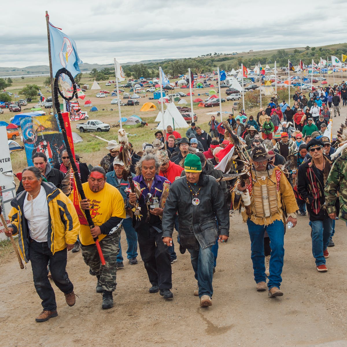 Color photograph of a large crowd of Dakota Access Pipeline protestors marching together through their encampment. Flags fly on poles along their path and tents, cars, tipis populate the grassy plain behind the marchers. Four of the Native American men in the foreground are carrying staffs and wearing face paint.
