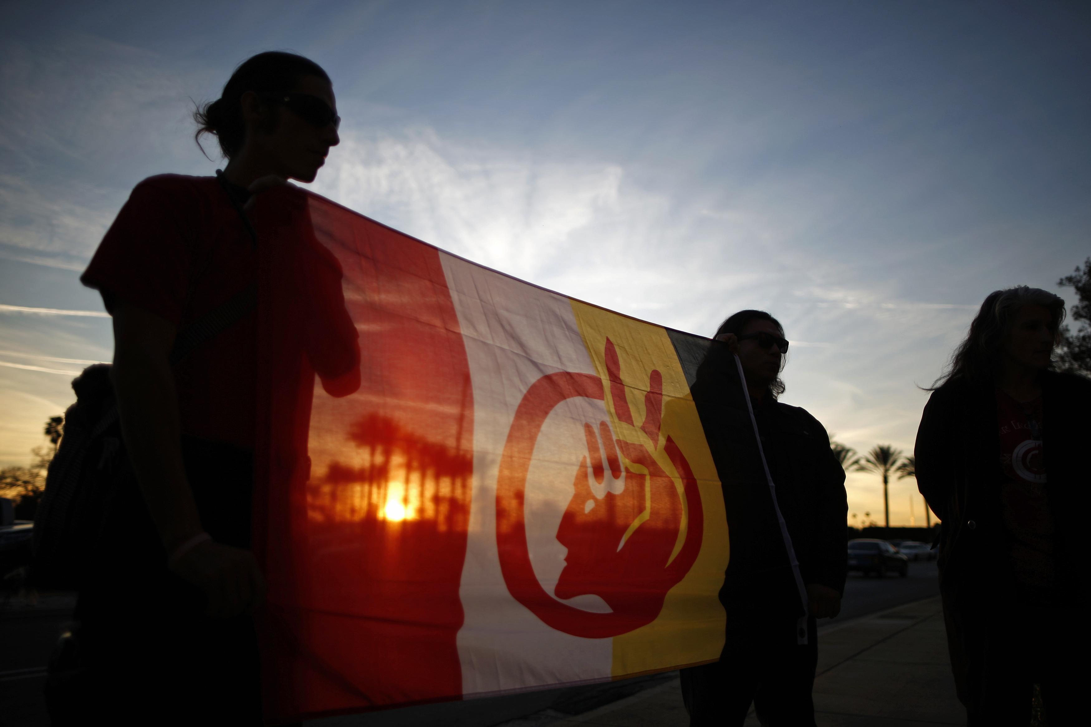 Color photograph of two women silhouetted against a setting sun holding an AIM flag between them. Another woman is slightly out of frame on the right. 