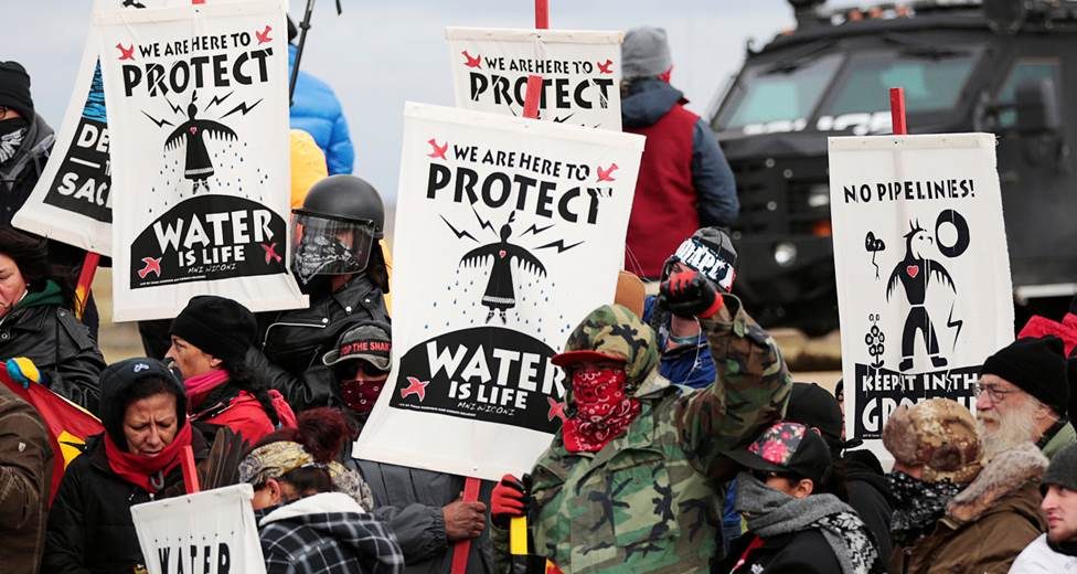 Color photograph of a group of Dakota Access Pipeline protesters consisting of members of the Standing Rock Sioux tribe, supporters from other tribes and environmentalists dressed in winter outdoor gear standing outside near the Standing Rock Reservation and the pipeline route by Saint Anthony, North Dakota. Many of the protestors are holding signs. Several are holding a sign that reads 'WE ARE HERE TO/PROTECT/WATER/ IS LIFE' printed in black and white. Another sign reads 'NO PIPELINES/KEEP IT IN THE GROUND'. Both signs show Native iconography. There is a black surveillance van in the background of the image. Photo by Reuters.
