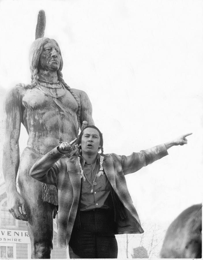 Black and white photograph of Russell Means speaking with arms raised and index fingers stretched pointing to the right. He stands in front of a large statue of Chief Massoit.