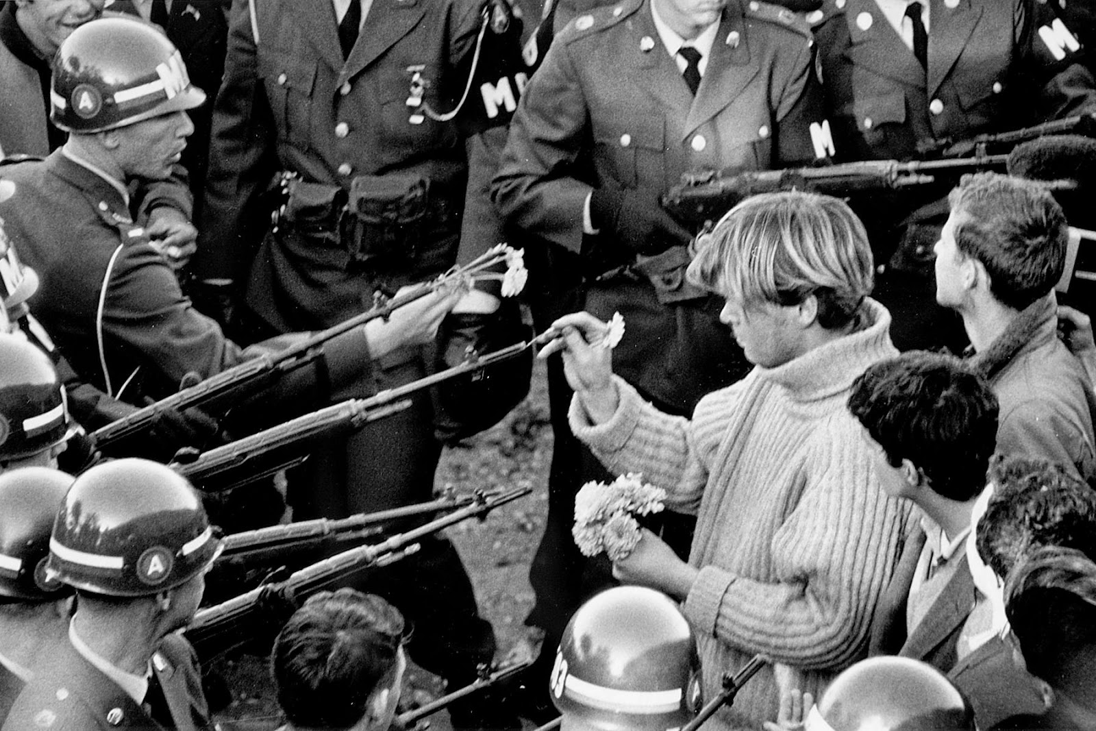 Black and white photograph featuring a group of White soldiers holding rifles, surrounding and pointed toward a group of young people, casually dressed. A young White man holds flowers in one hand as he places the stem of a single flower into the barrel of a soldier’s gun.