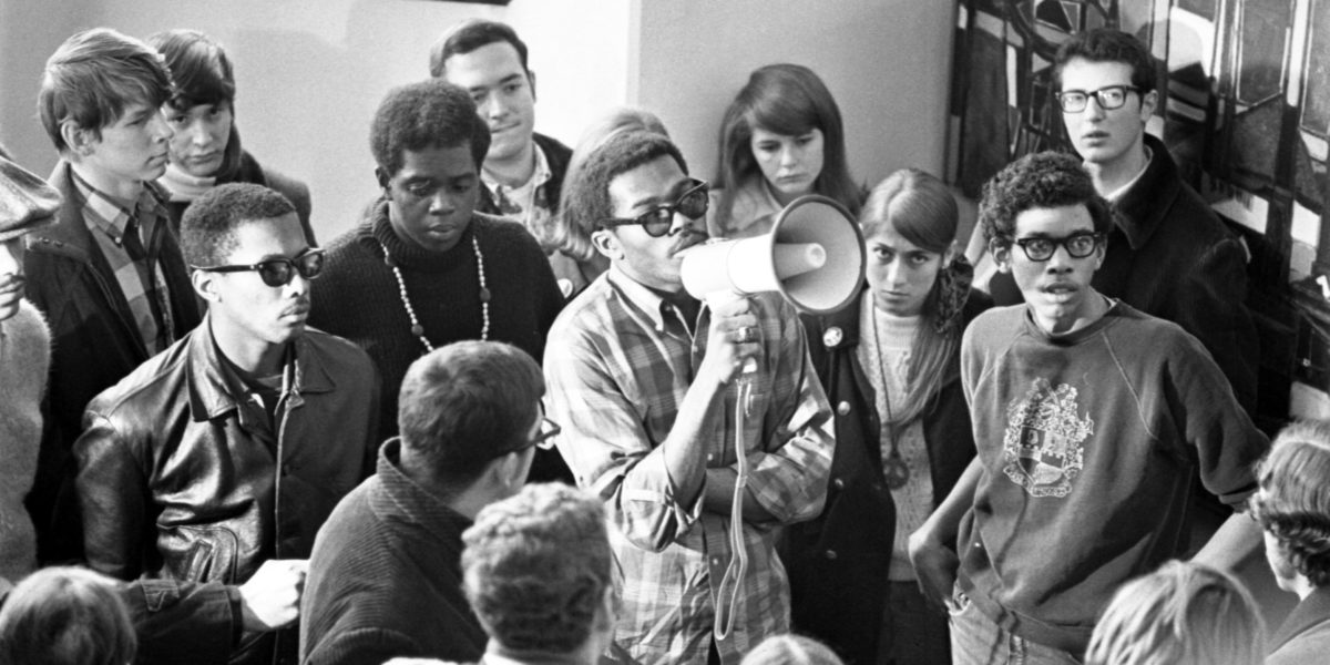 Black and white photograph showing a group of fourteen young Black and White men and women at a protest. There is a Black man holding a megaphone in the center of the group.