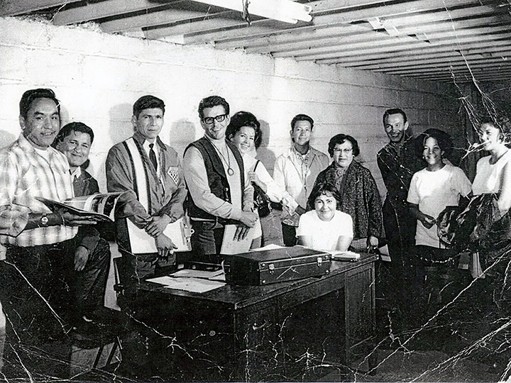 Black and white photograph showing the gathering of eleven young Native American men and women at the first official meeting of the American Indian Movement in 1968. The group is gathered around a desk in the basement of a building. Russell Means is in the photo.