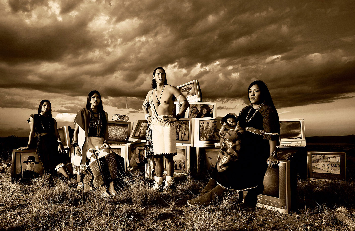  Sepia toned photograph of four adult Native Americans and a baby in traditional clothing posed in front of a series of television sets on a grassy landscape in the foreground; the television sets show images of Native Americans as represented on television or film as well as photo-documentary images that include raising the flag on Iwo Jima, a nuclear test site mushroom cloud, and an image of graffiti from the occupation of Alcatraz by Indians of All Tribes (IOAT); the background shows a dark horizon line and an abundance of clouds overhead.