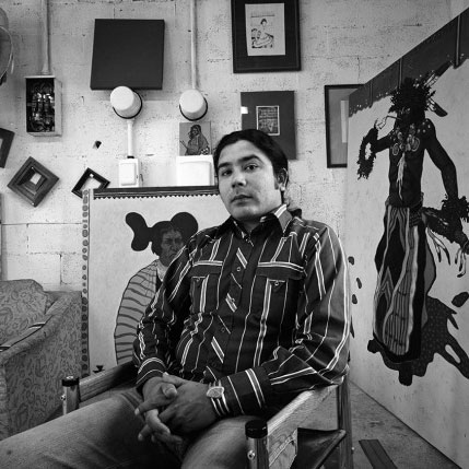 Black and white photograph of artist T.C. Cannon sitting in his studio surrounded by works of art.