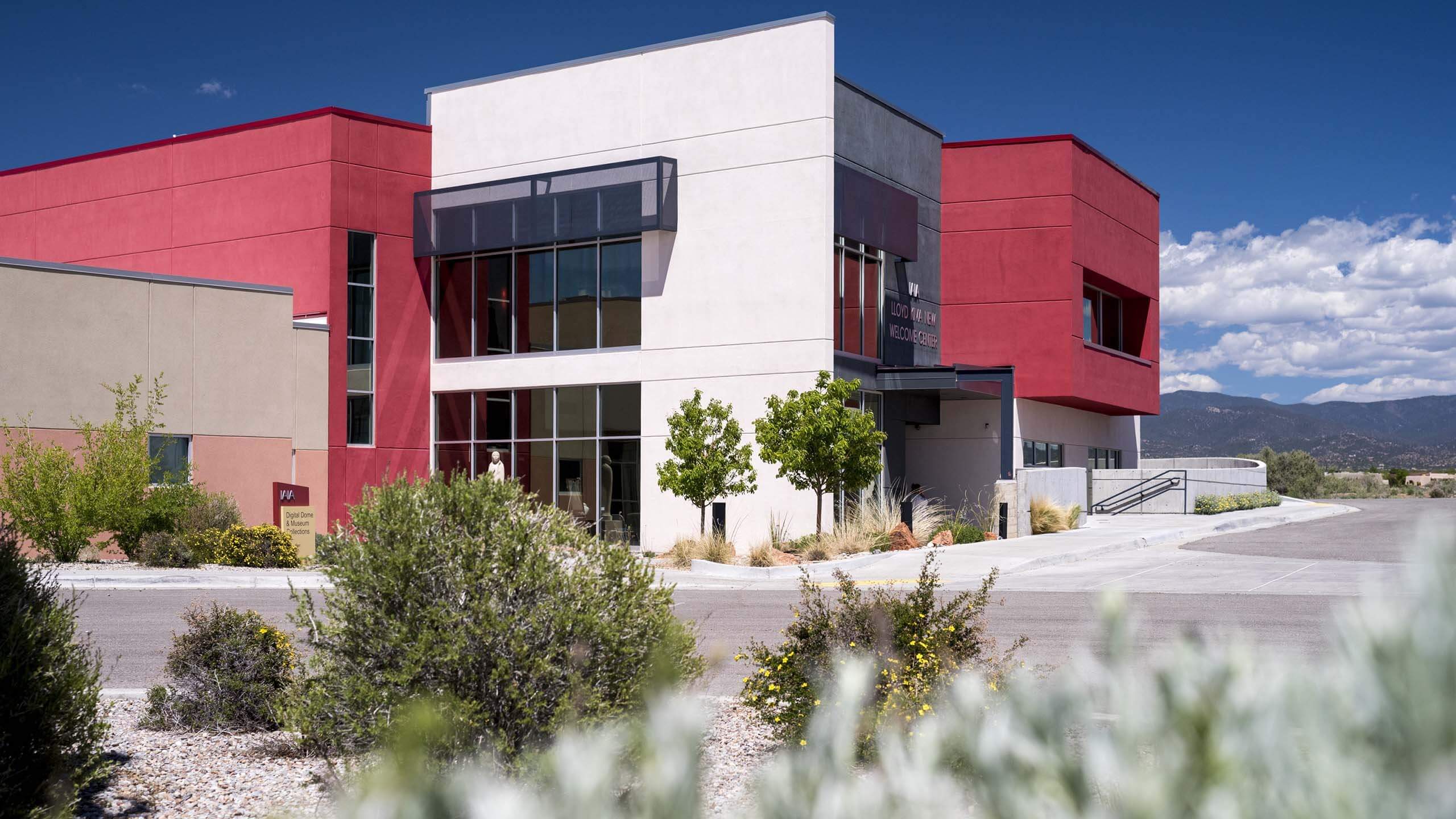 Color photograph of the exterior of the Institute of American Indian Arts (IAIA) in Santa Fe, New Mexico. The contemporary building consists of several angled surfaces with many windows; the colors are predominantly red, white, and black/gray.