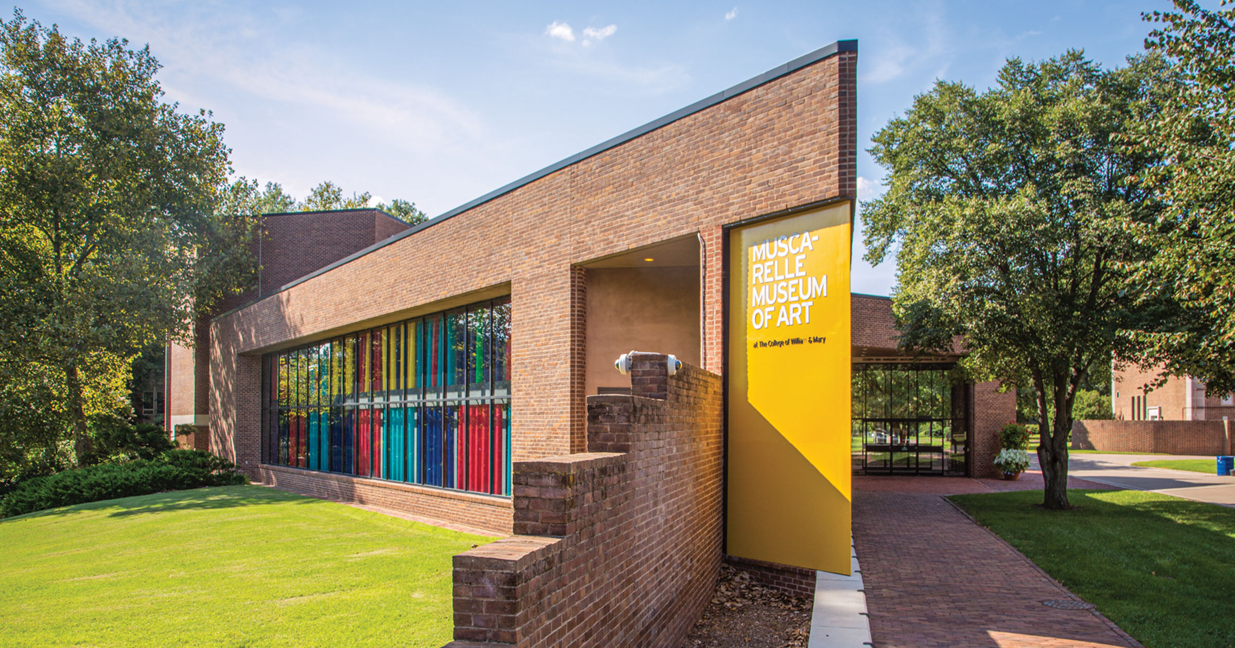 Color photograph of the exterior of the Muscarelle Museum of Art at William & Mary in Williamsburg, Virginia. Building is red brick and one exterior wall features Sunlight Sonata, a solar light wall consisting of two levels of yellow, turquoise, cobalt, and red tubes created by artist Gene Davis for the museum.