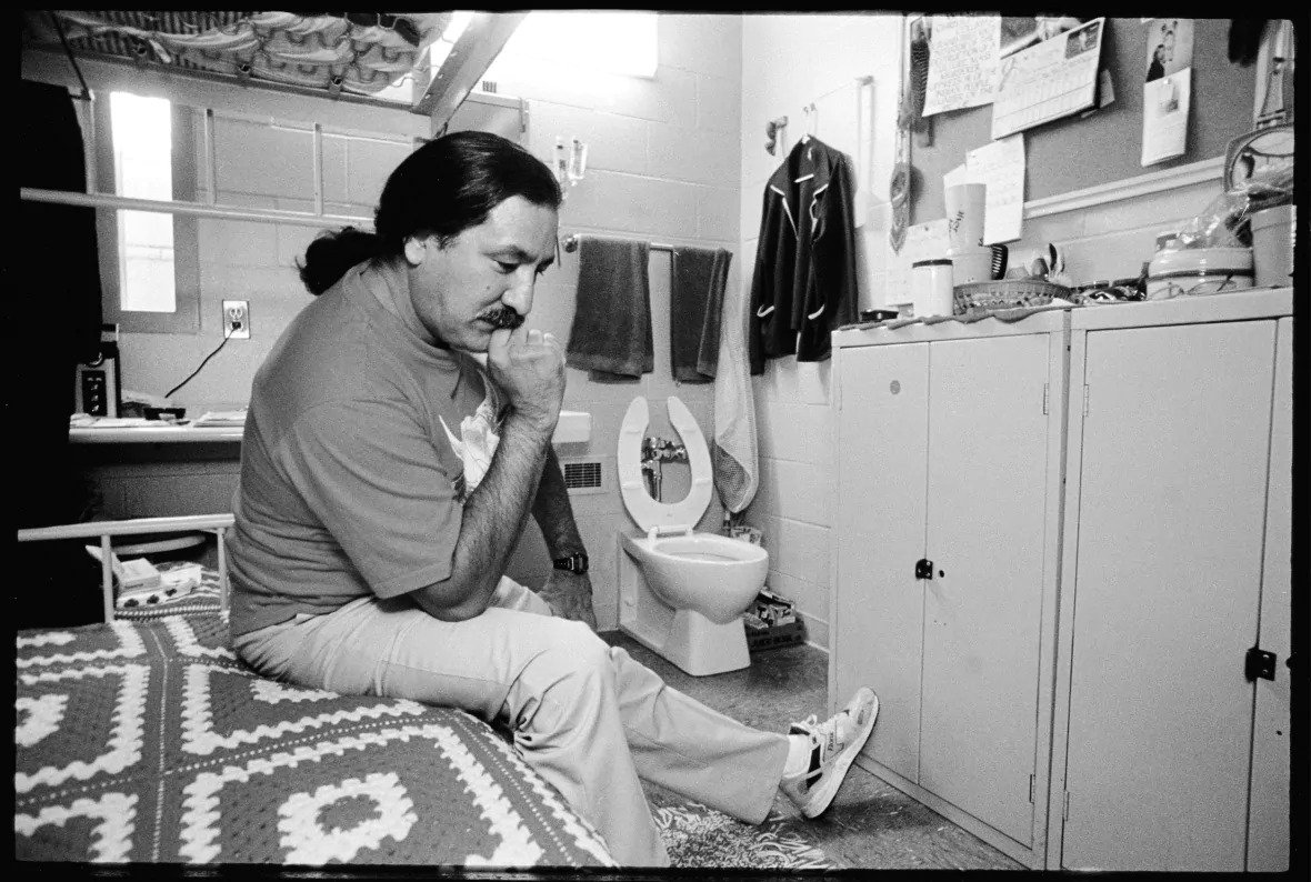 Black and white photograph of Leonard Peltier sitting on his prison bed. On the opposite wall there are personal effects hung on a bulletin board and on top of cabinets. Peltier appears to be thinking while looking at the floor.