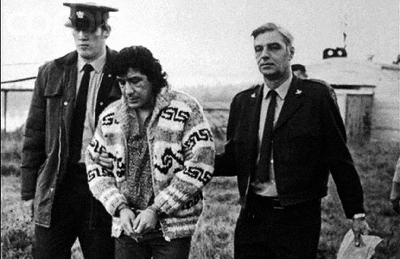 Black and white photograph of two White male law enforcement personnel grasping the handcuffed Leonard Peltier’s arms as he is taken into custody.